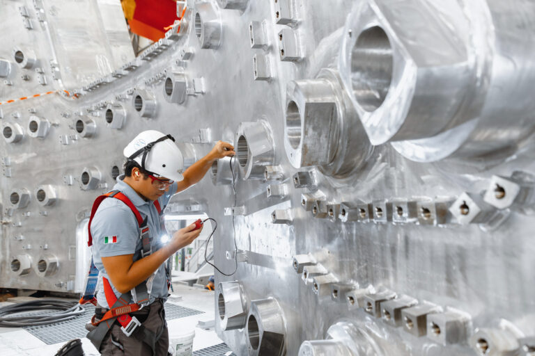 A construction worker in a hard hat regarding a measuring device in his hand with a background of stainless steel wall with openings.