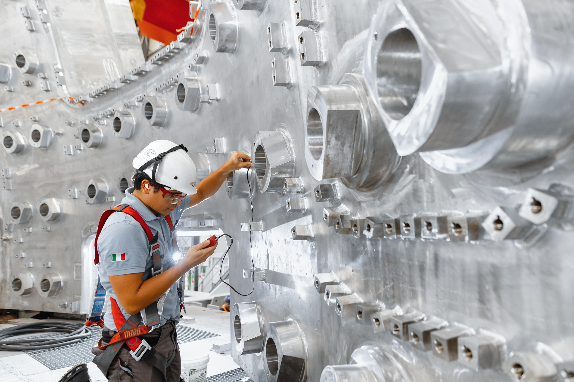 A construction worker in a hard hat regarding a measuring device in his hand with a background of stainless steel wall with openings.