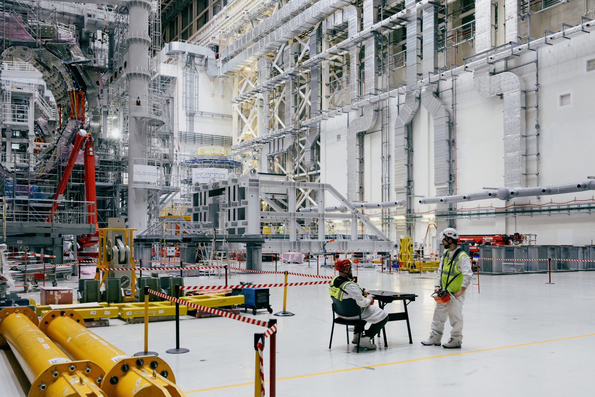 Two workers in the foreground give a sense of scale to a vast hall lined with ventilation pipes and construction devices.