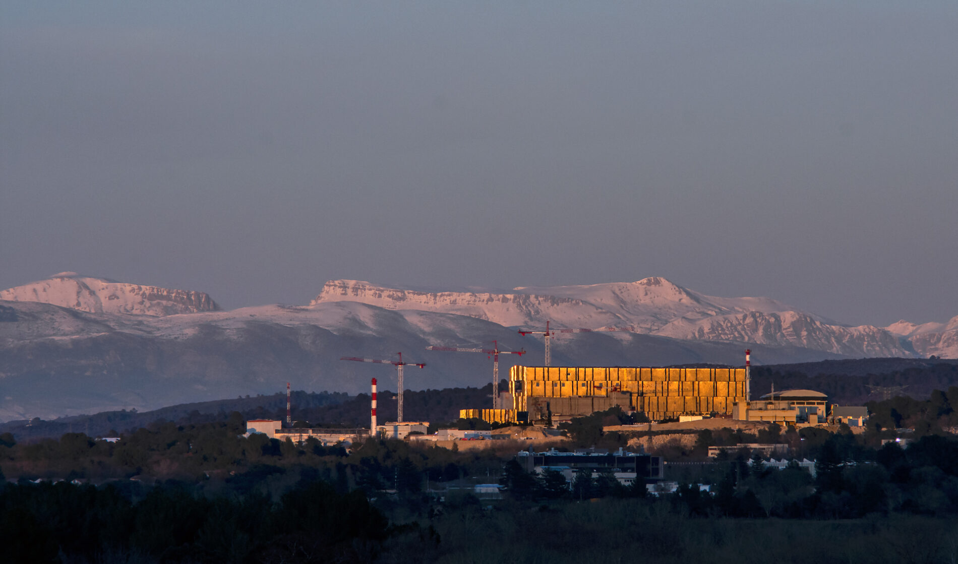 A large industrial complex with several construction cranes rising from it. Snow-capped mountains fill the background.