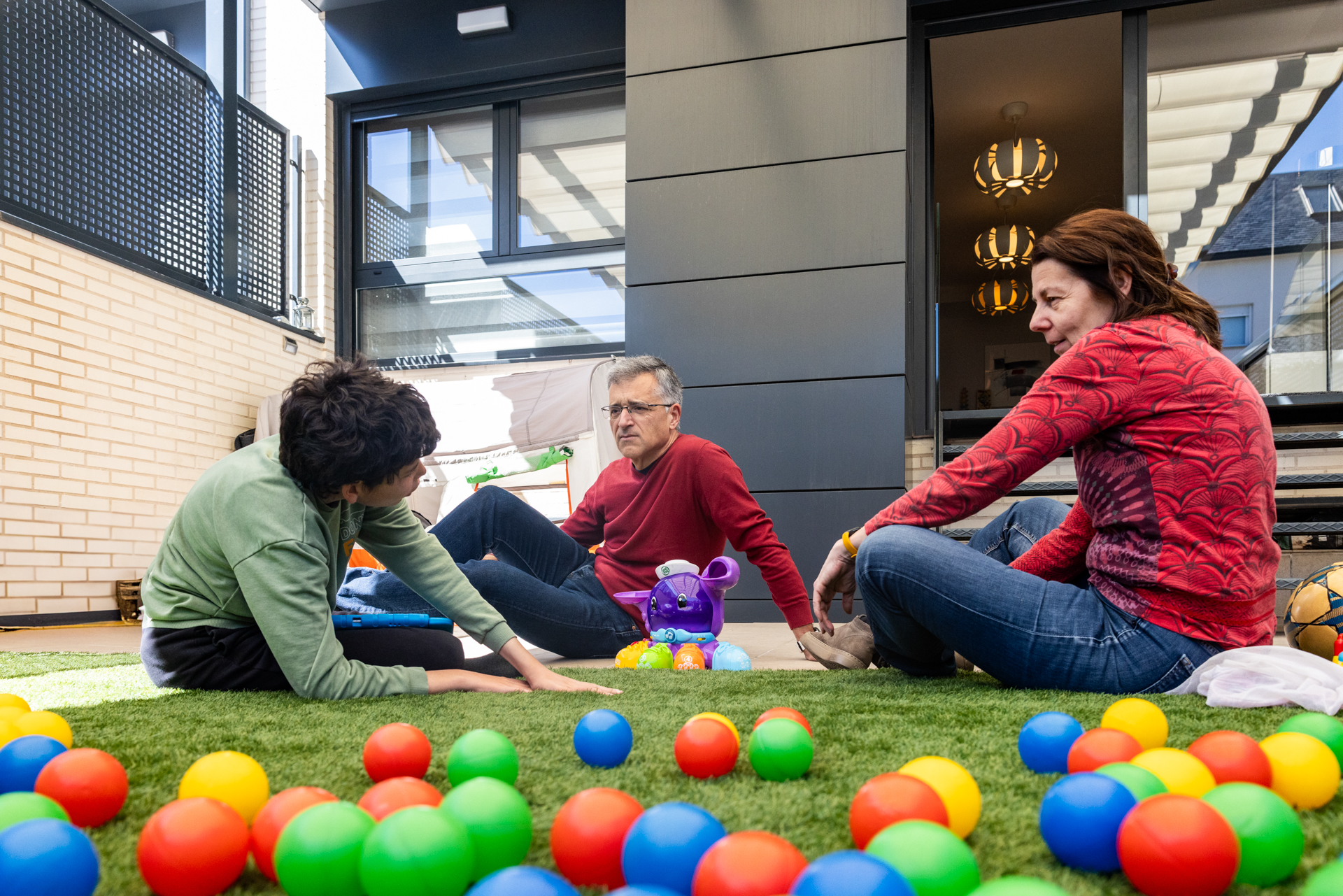 A man, woman and child sit in a garden surrounded by colorful plastic play balls.