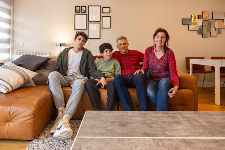 A family of four at home sit on a sofa looking at the camera.