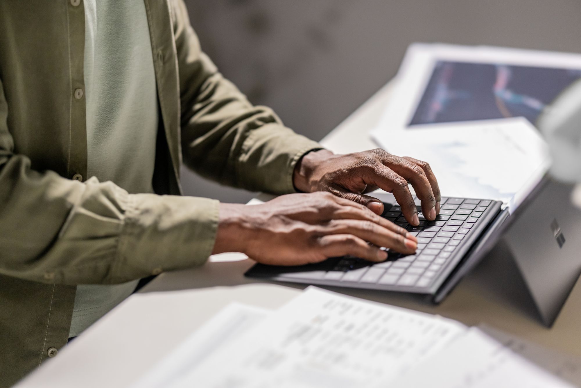 An African man types on a Surface laptop that is set on a desk, with documents opened alongside the laptop.