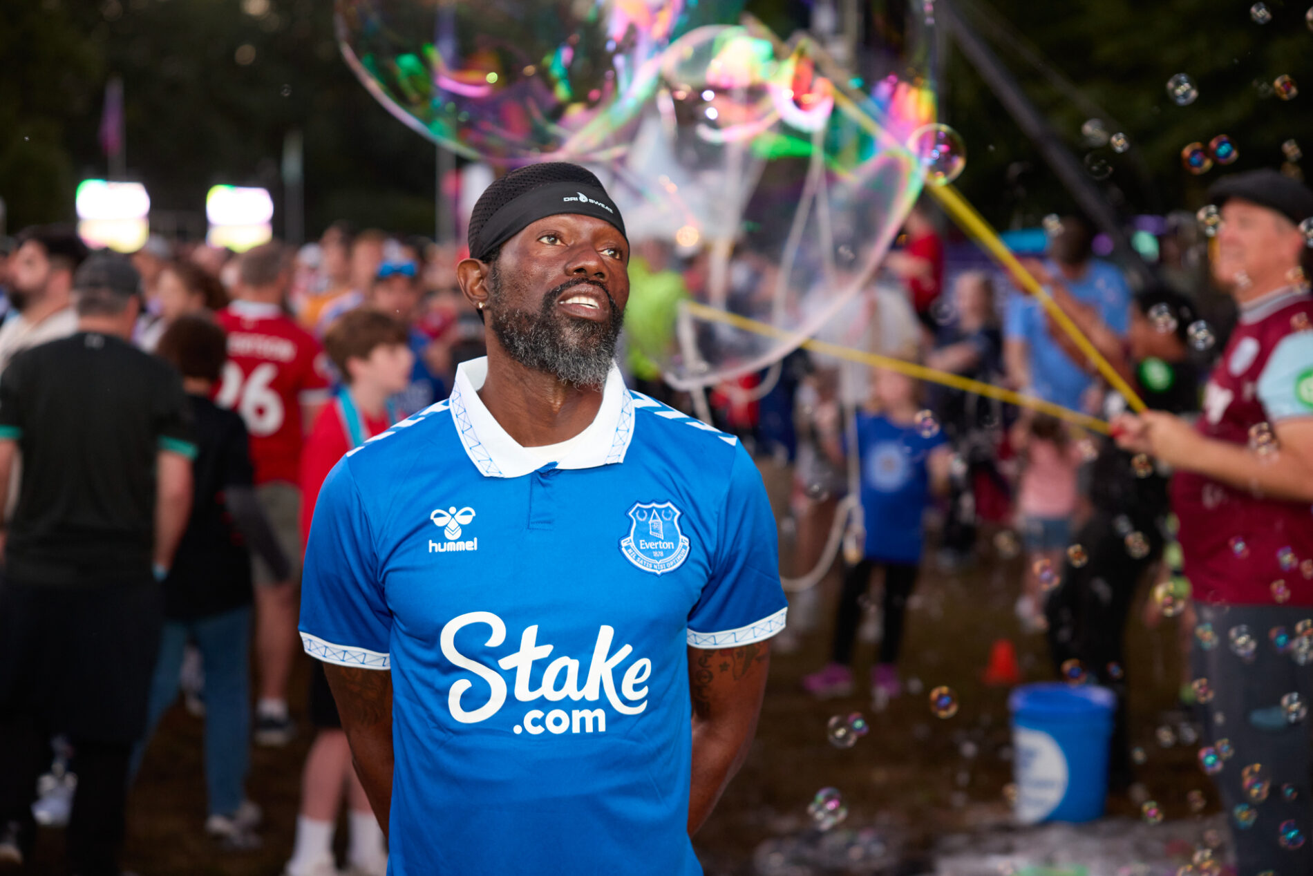 A man wearing a blue Everton jersey looks forward while a large bubble is blown behind him.